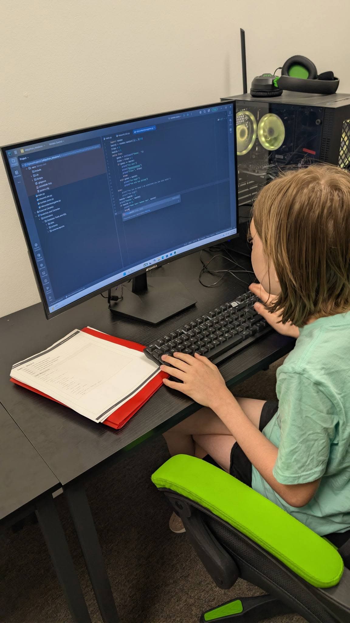 A child absorbed in real code on a computer screen, hand on the keyboard, with a project folder open on the desk beside them.