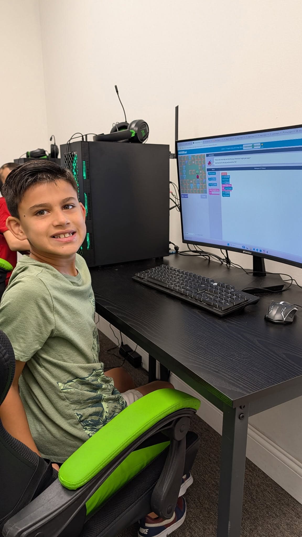 A boy smiling at the camera beside a computer showing a beginner block-coding puzzle, in a classroom with a keyboard and mouse in front of him.