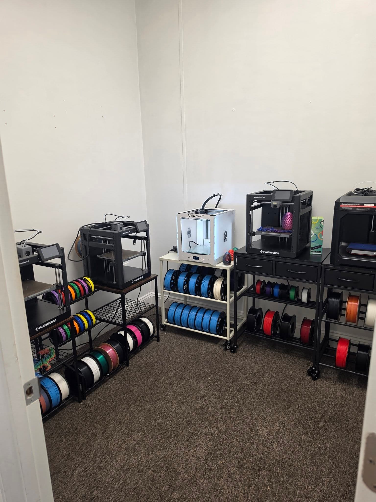 Looking through the open doorway into the 3D printing room, with several printers and racks of colored filament along the back wall.