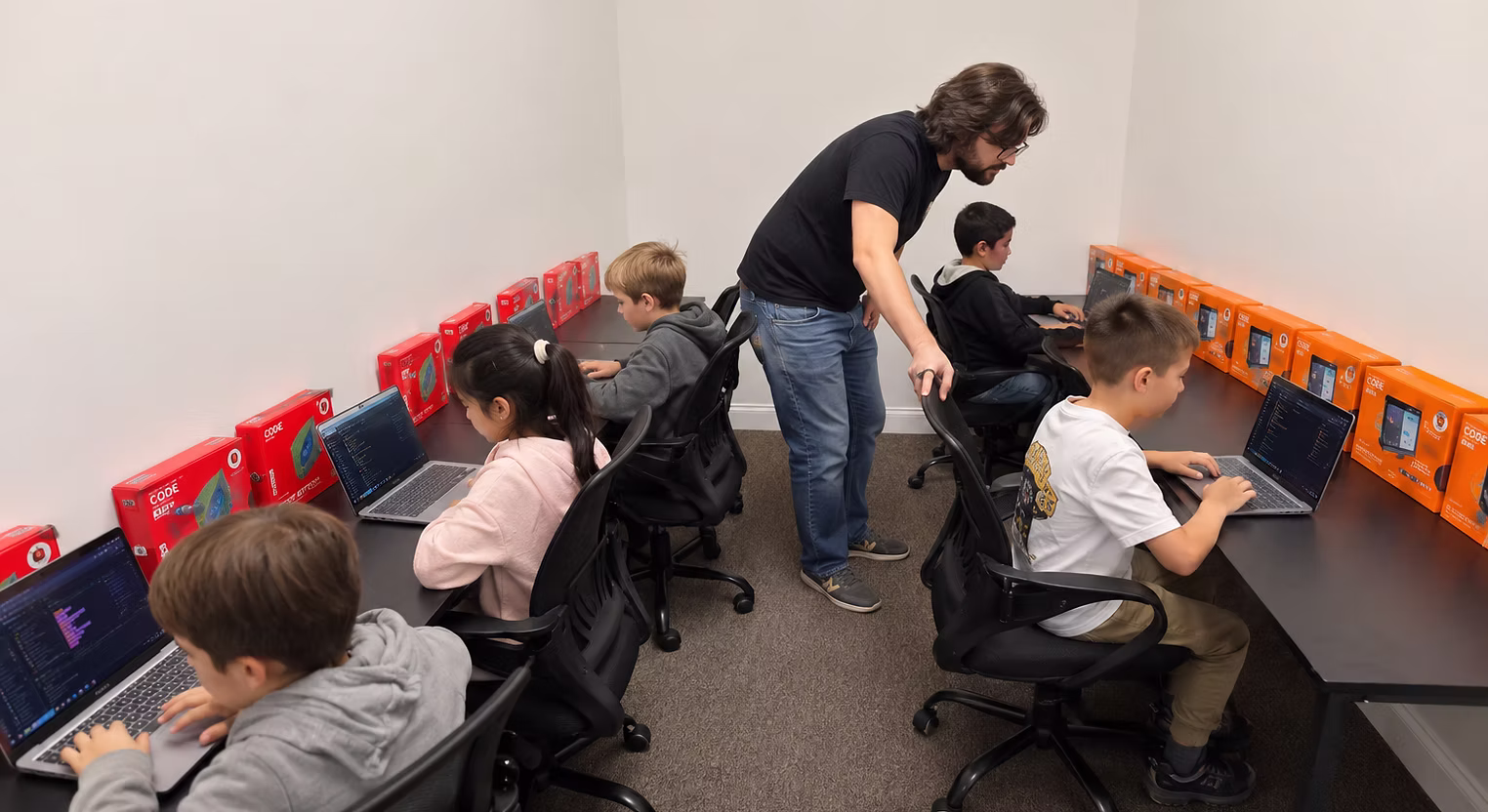 The same RunTheCode coding room with students working at laptops while an adult moves through the room.