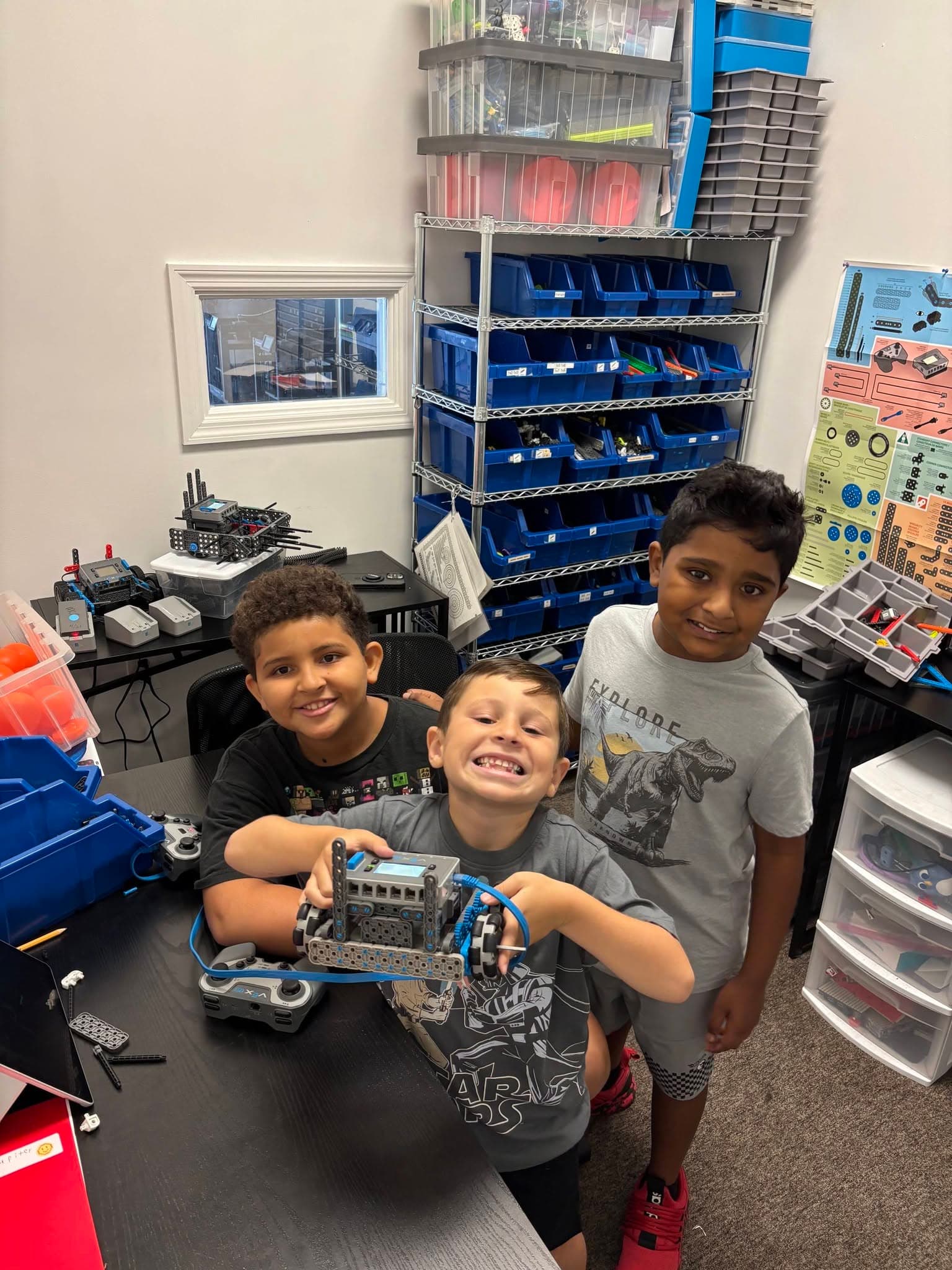 Three kids smiling together while holding a robot they built, with robotics storage and parts visible in the background.