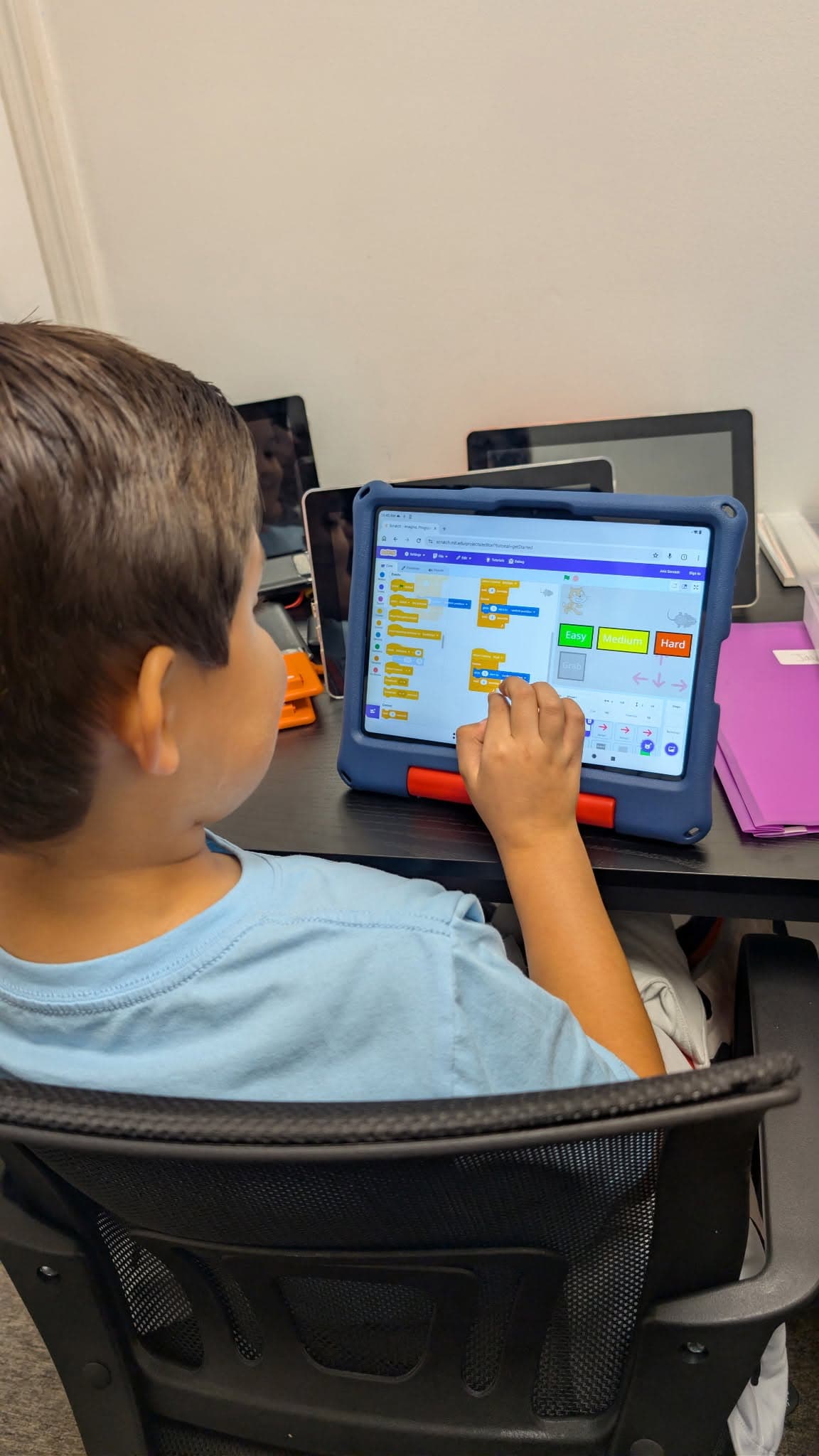 A student dragging code blocks on a tablet to build a game, with a character sprite and difficulty-level buttons visible on screen.