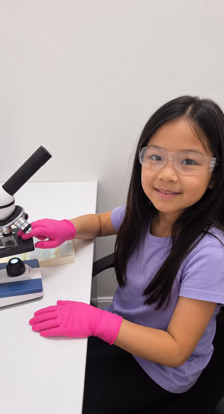 A student in safety glasses and gloves smiling while using a microscope.