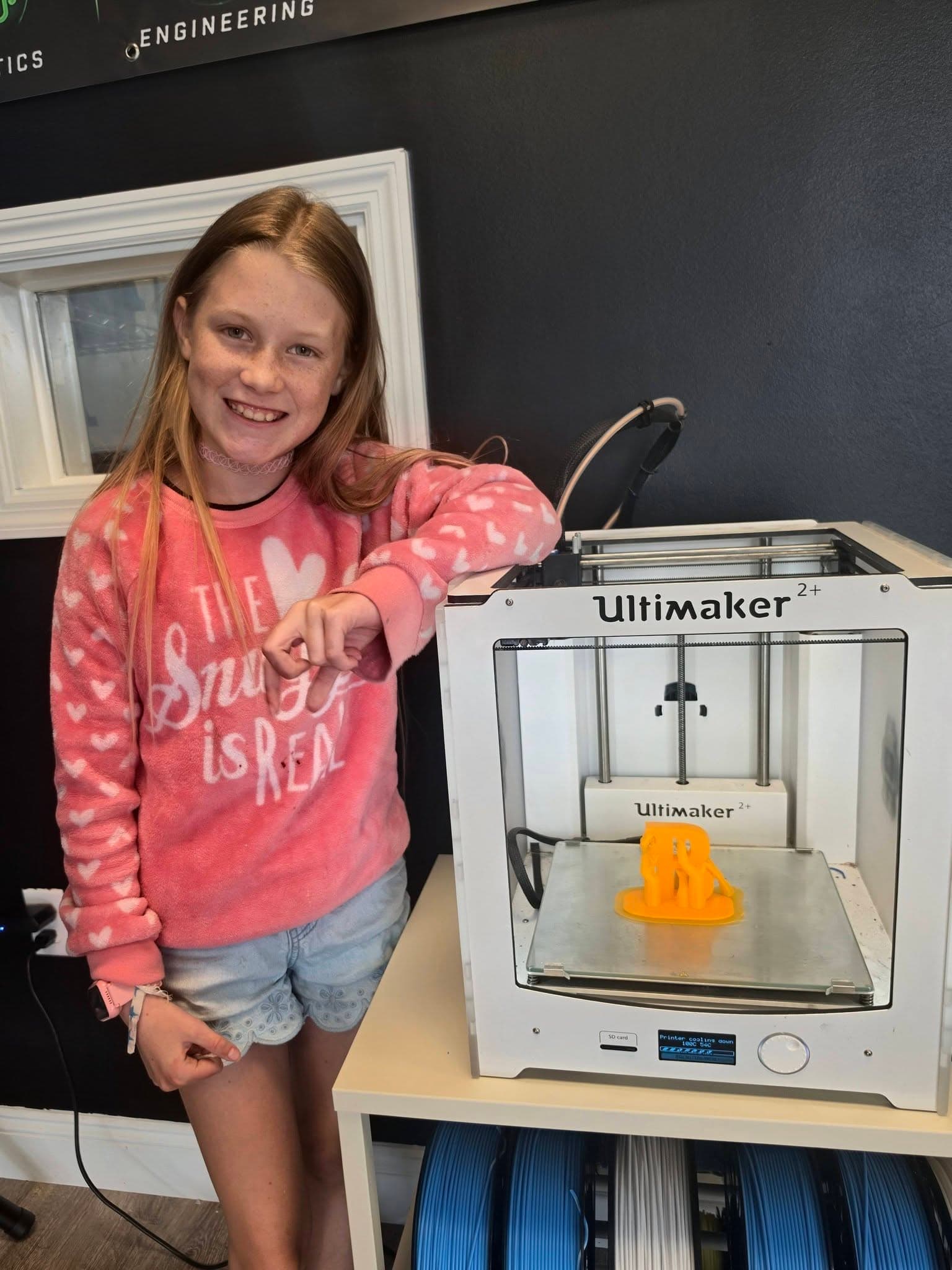 A student standing beside an Ultimaker 2+ 3D printer mid-print, in front of real Robotics and Engineering lab signage.